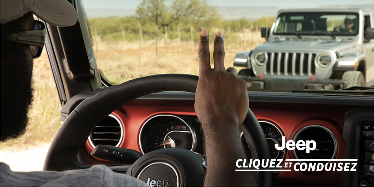 Cliquez et Conduisez avec Jeep. Une personne dans un Jeep Wrangler roulant sur une route de terre salue une autre personne qu’elle croise dans un Jeep Wrangler gris.
