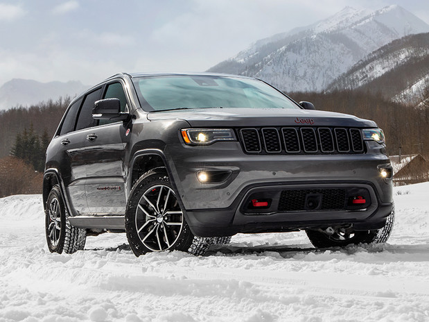 Jeep Grand Cherokee gris garé dans un vaste paysage enneigé avec des arbres, des maisons et des montagnes en arrière-plan.