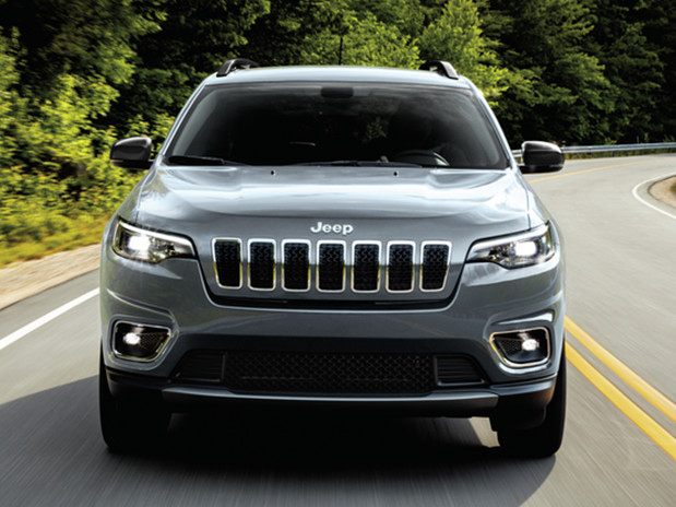 Front view of a silver Jeep Cheroke driving on the highway with a forest background.