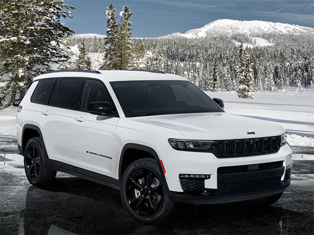 Side profile view of a white 2023 Jeep Grand Cherokee 4xe PHEV parked with snow covered trees and mountains in the background.