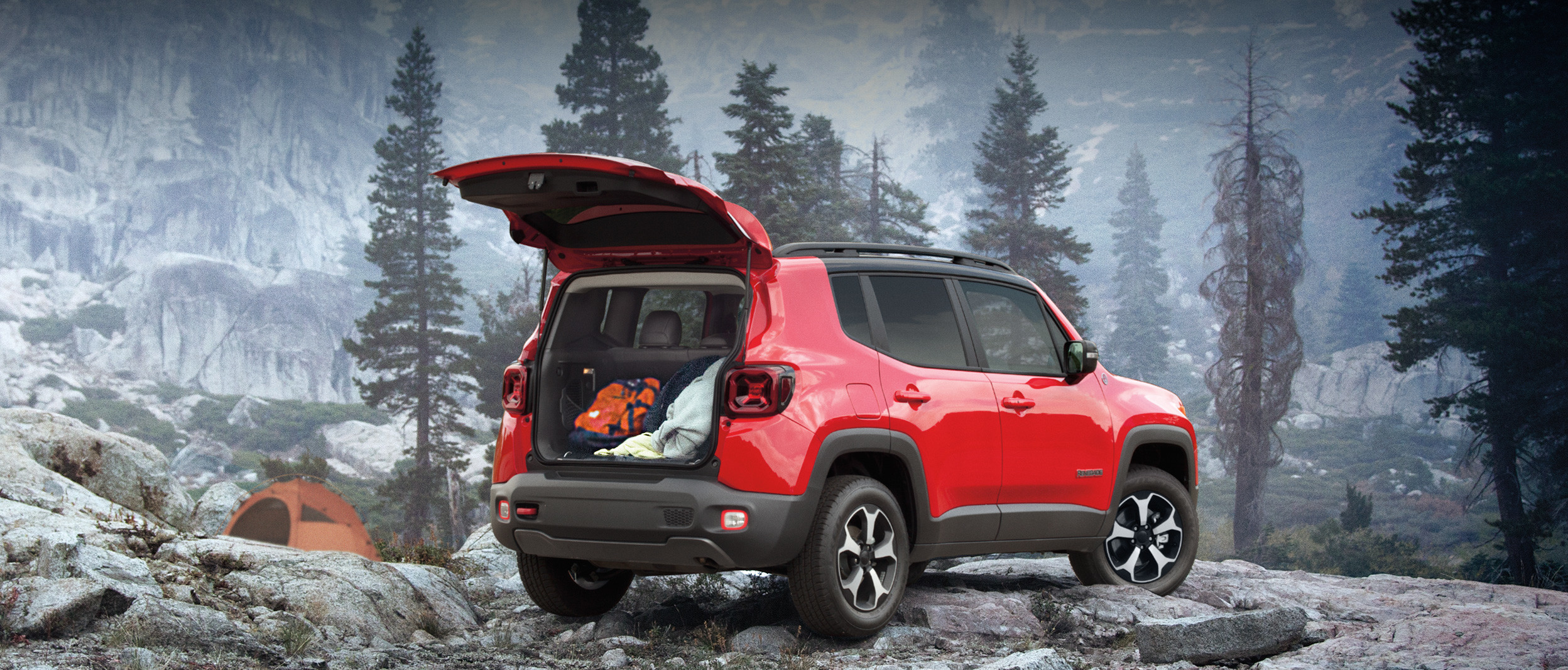 Vue arrière du Jeep Renegade 2020, illustré en rouge, stationné dans une forêt rocheuse à même les montagnes, près d’un homme qui prépare de la nourriture sur un réchaud de camping