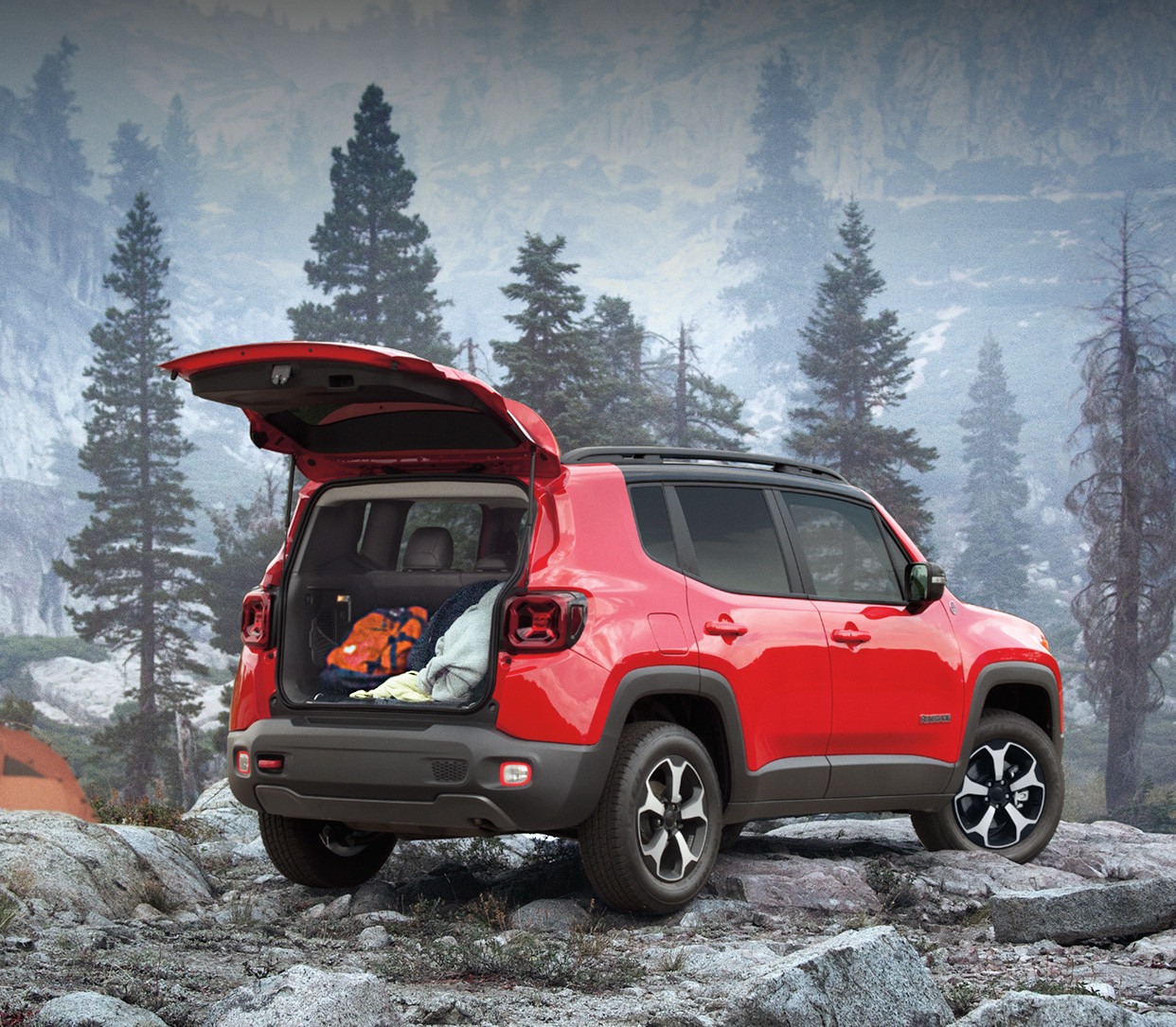 Vue arrière du Jeep Renegade 2020, illustré en rouge, stationné dans une forêt rocheuse à même les montagnes, près d’un homme qui prépare de la nourriture sur un réchaud de camping