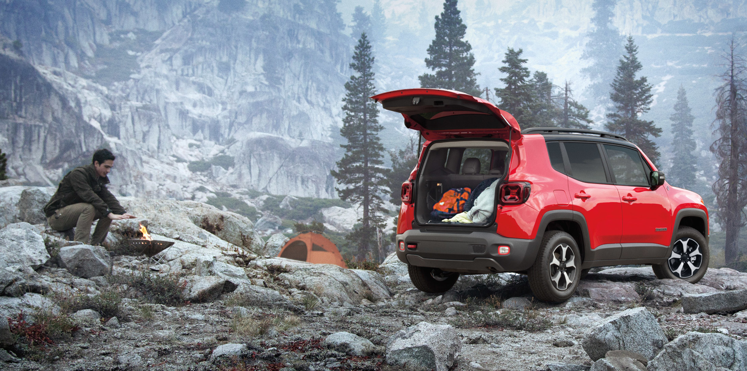 Red Jeep Renegade parked with the trunk open, while a man on a camping trip warms his hands by a fire nearby.