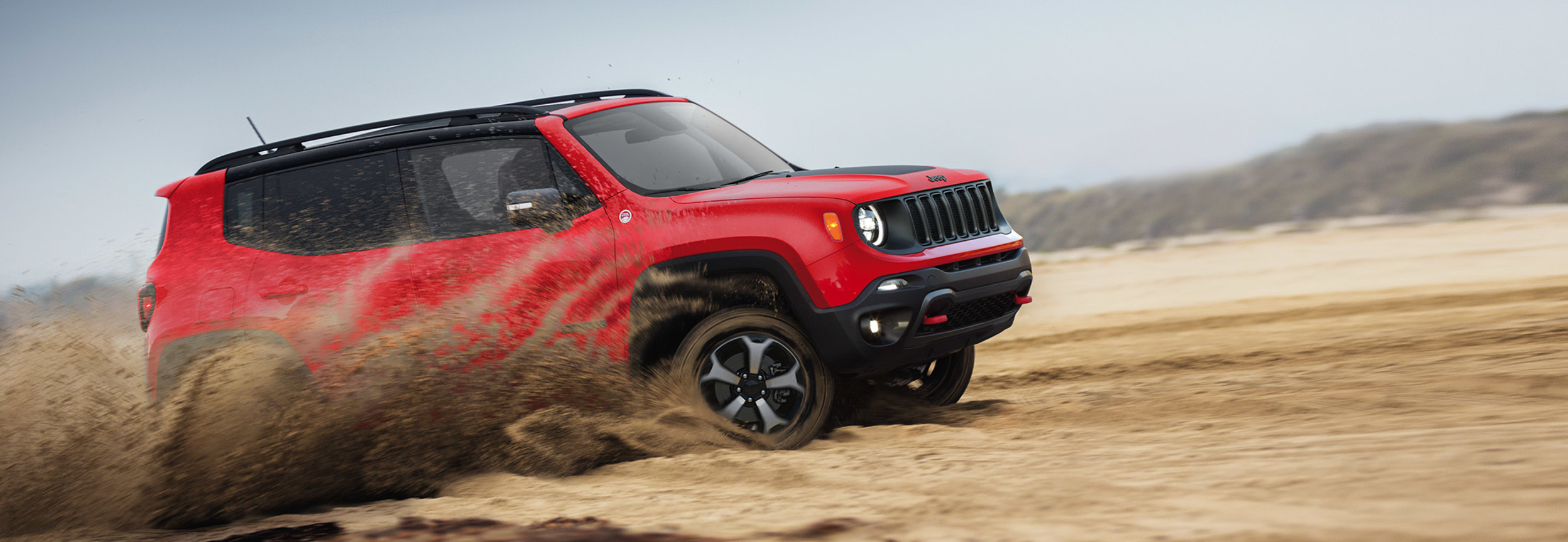 Jeep Renegade off-roads through a desert landscape, kicking up sand.