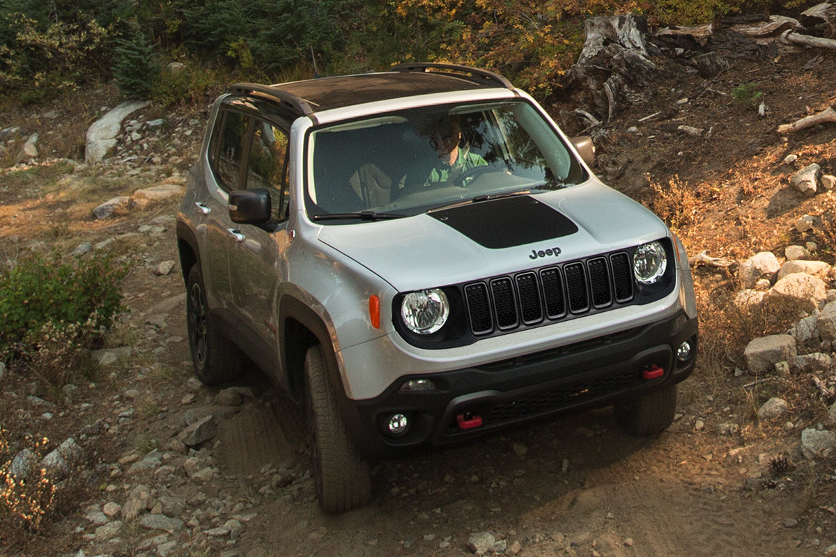 Grey Jeep Renegade off-roads on a trail with trees and rocky hills in the background.