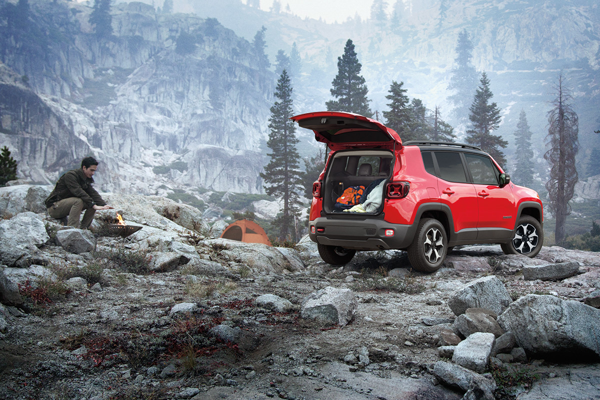 A landscape view of a mountain with a man starting a fire with a red 2022 Jeep Renegade parked to the side with the trunk open.