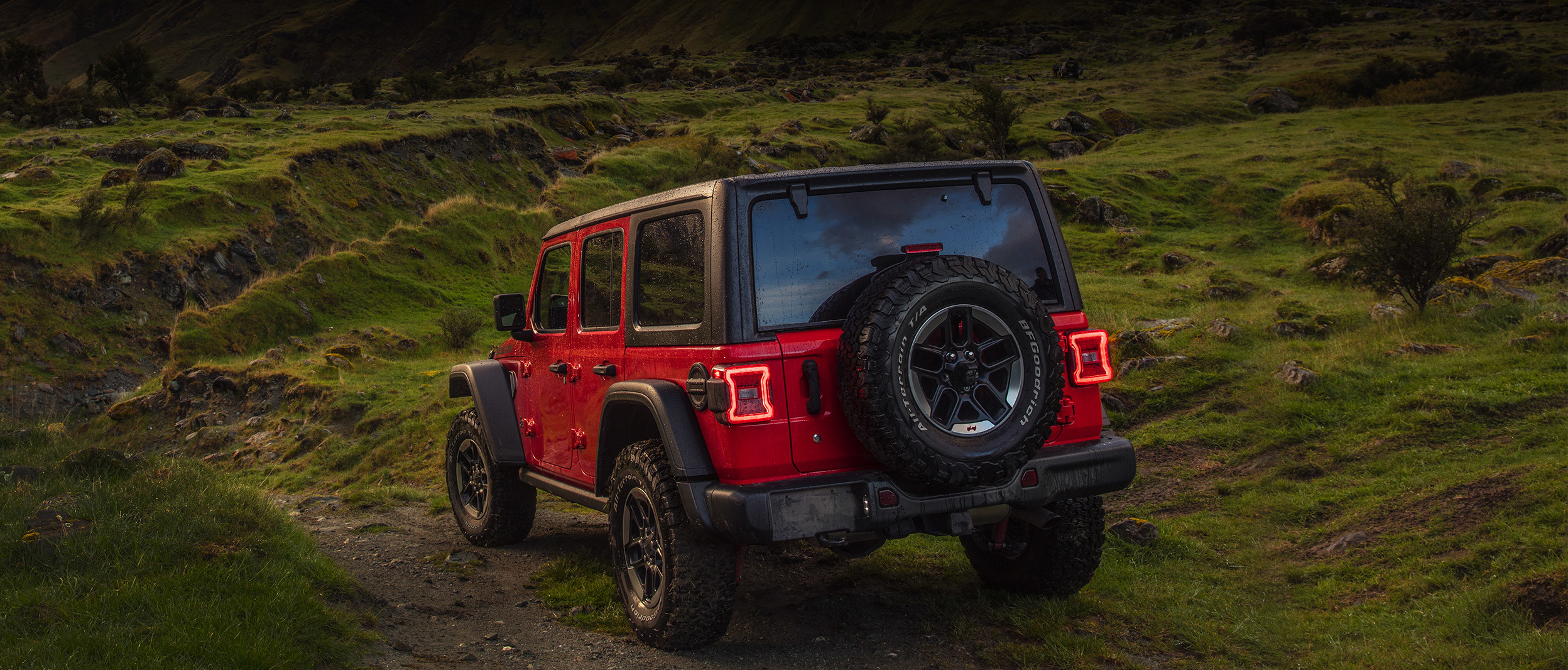 Rear quarter view of the red 2020 Jeep Wrangler parked on a dirt road facing mountains during sunset