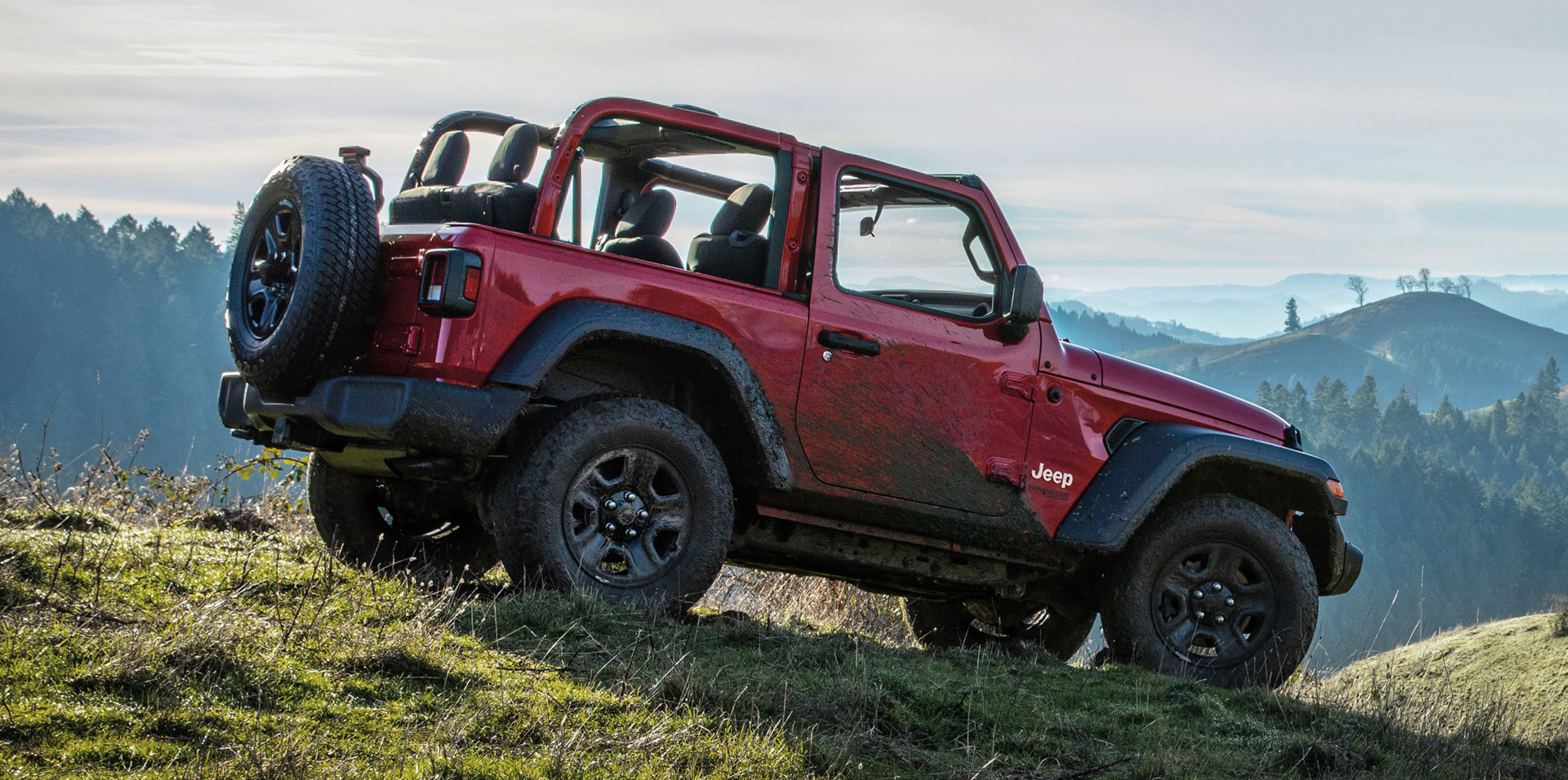 Jeep Wrangler rouge couvert de boue garé sur une colline verdoyante avec des montagnes en arrière-plan.