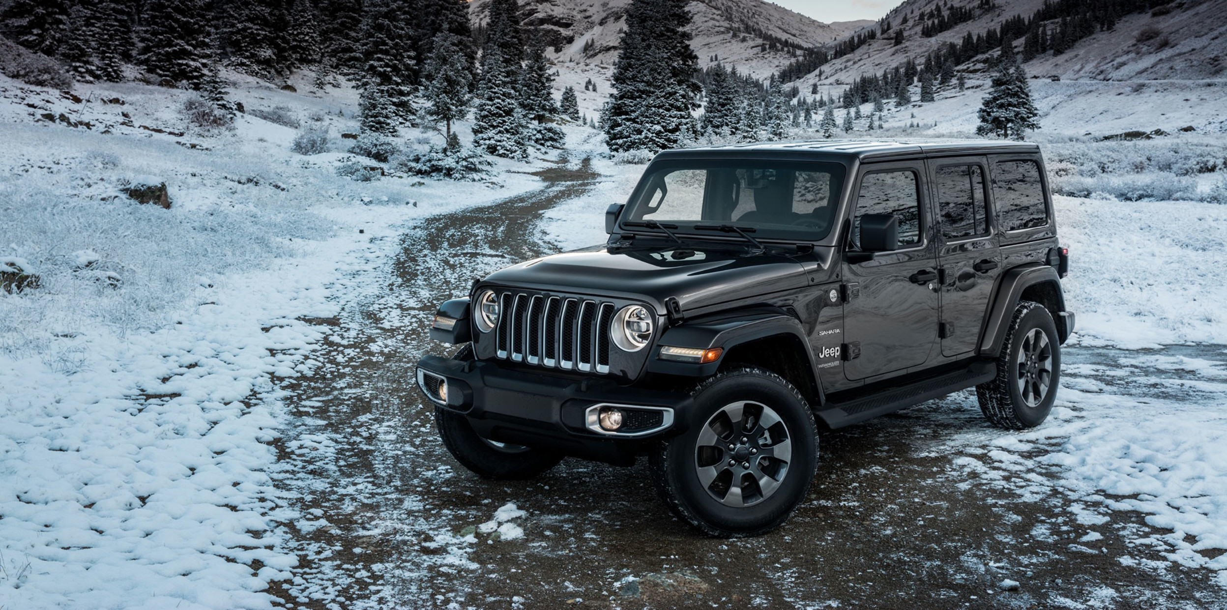 Profile view of a black 2023 Jeep Wrangler parked on a dirt path covered in snow and mountains in the background.