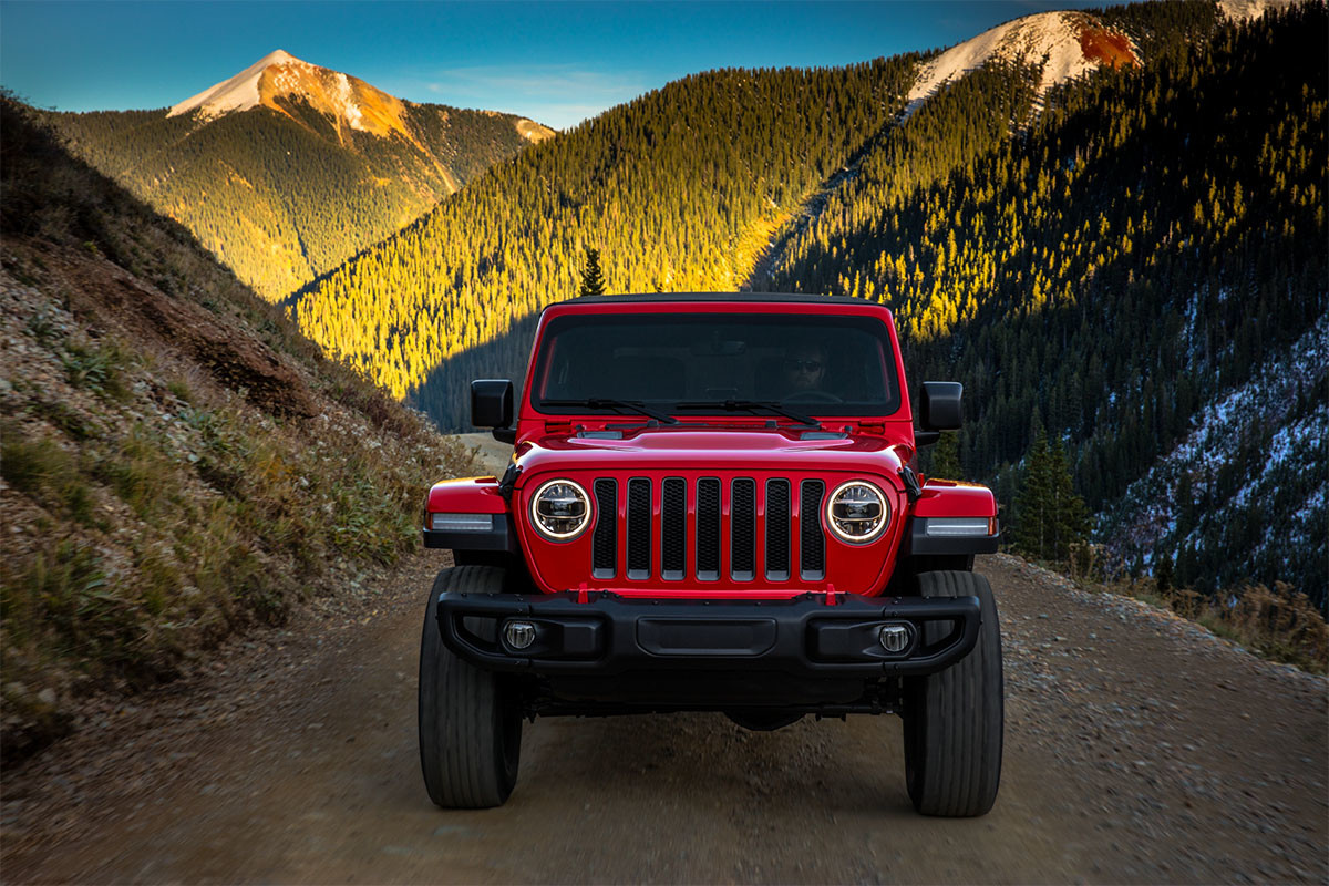 Front facing view of a red 2023 Jeep Wrangler parked on top of a gravel road with tree covered mountains shown behind.