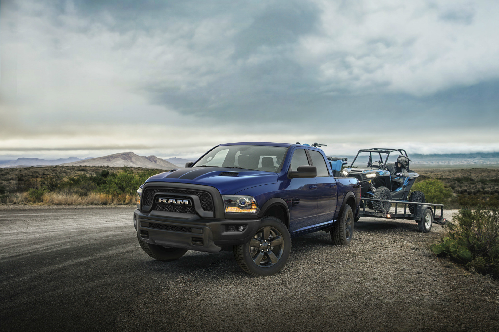 Vue de face du Ram 1500 Classic 2019, illustré en bleu, remorquant un véhicule tout-terrain sur une route de terre
