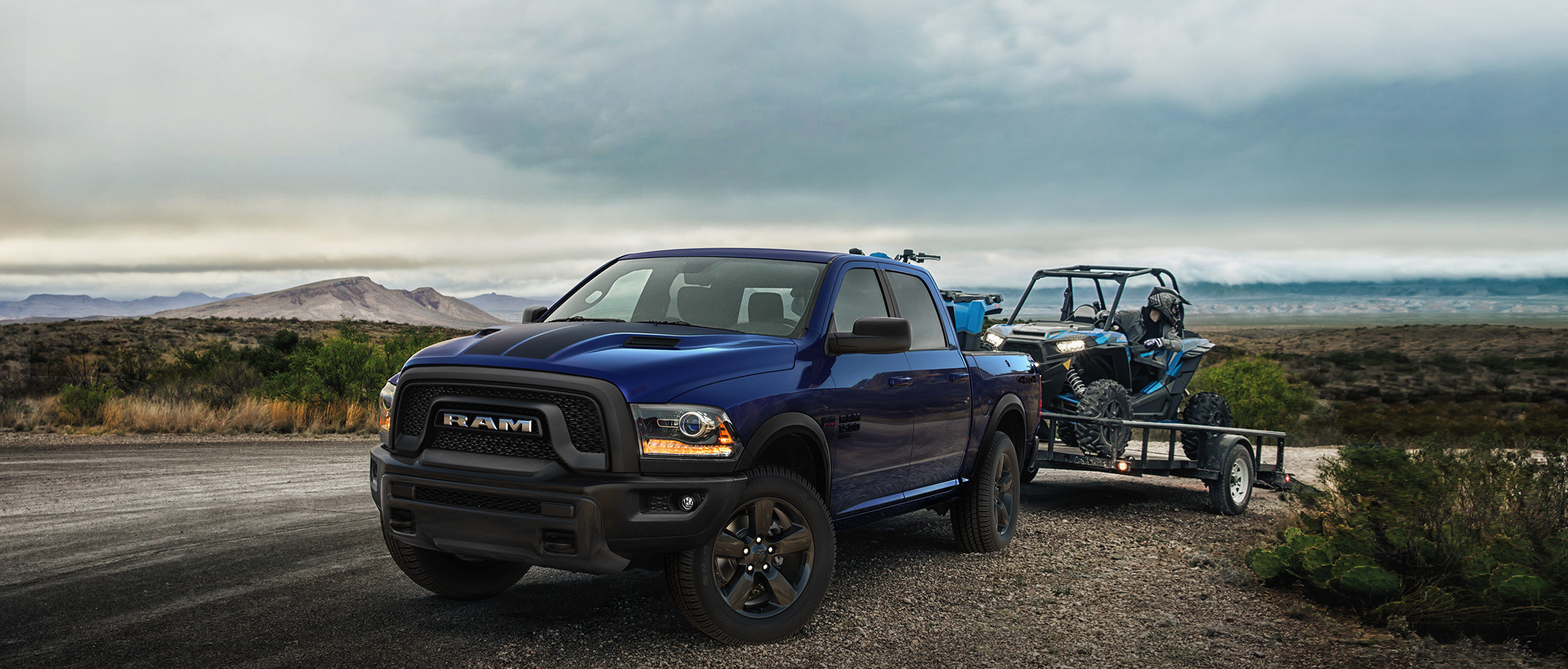 Blue 2020 Ram 1500 Classic, parked on a gravel road, towing a blue ATV on a utility trailer by the mountains.