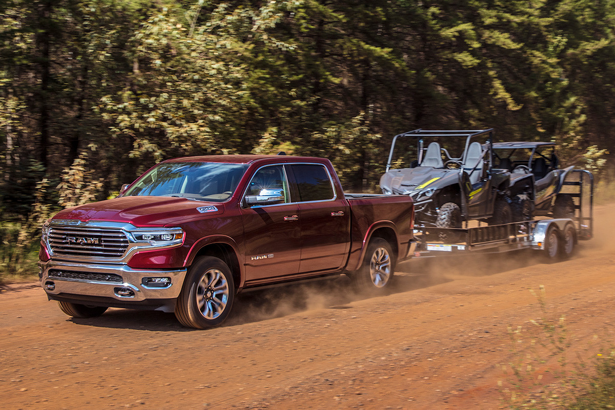 Red 2019 Ram 1500 DT being driven down a dirt road, with trees in the background, while towing two grey ATVs on a utility trailer.