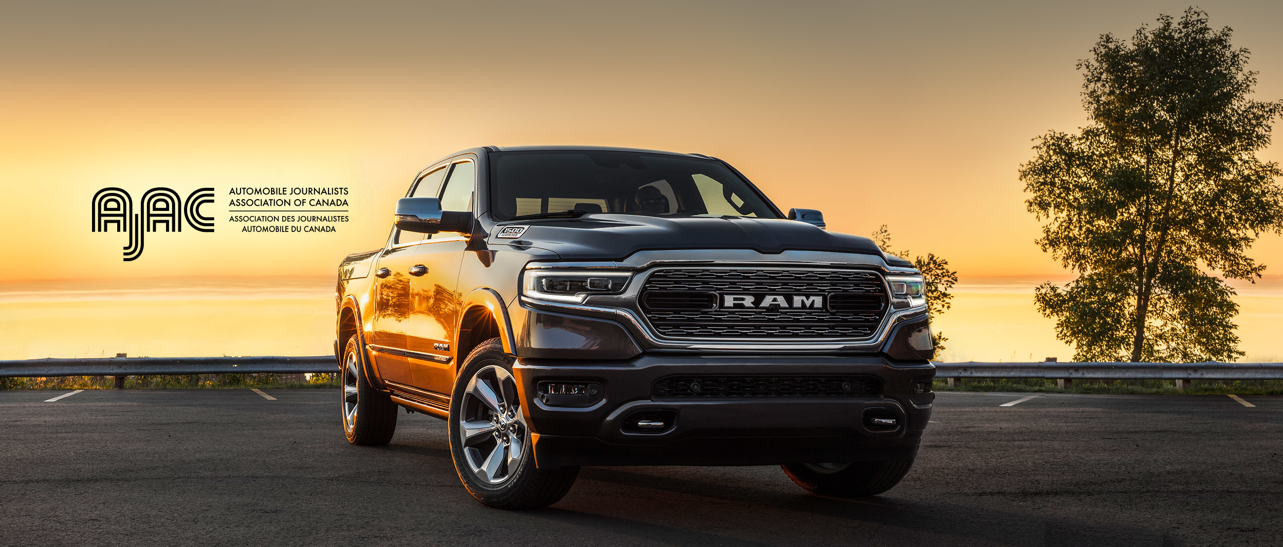 Front view of the 2020 Ram 1500 in black parked in front of the lake at sunset with the AJAC logo on top.