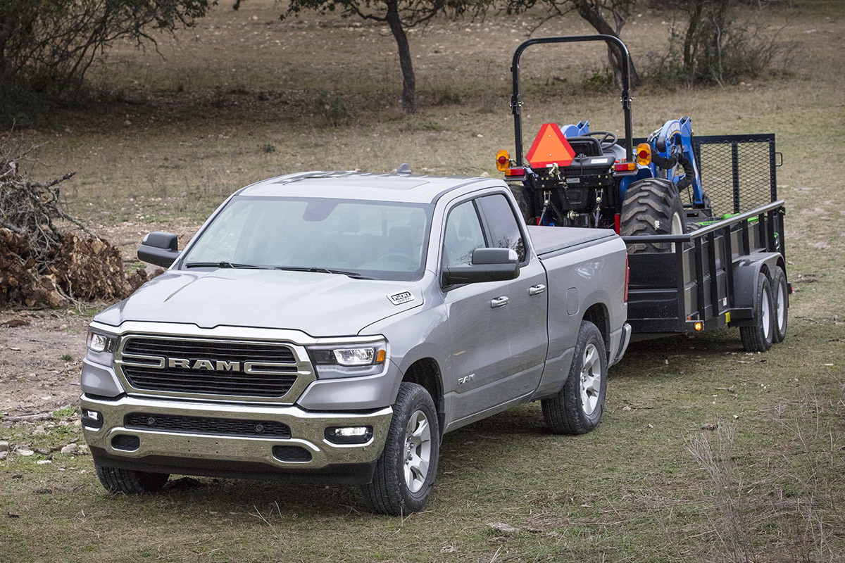 Vue avant d’un Ram 1500 DT 2021 gris acier métallisé tirant une remorque chargée d’un tracteur compact dans un champ.