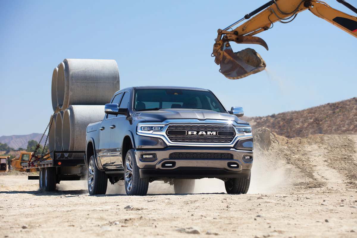 Vue d’un Ram 1500 DT 2021 gris acier métallisé tirant une remorque chargée de cylindres en ciment à côté d’une excavatrice sur un chantier de construction.