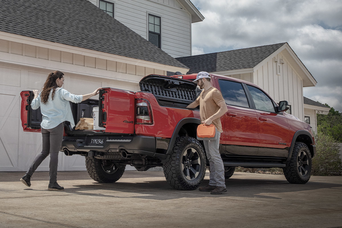 Vue arrière d’une femme ouvrant les portes à battants divisés 60/40 du hayon multifonction livrable en option sur un Ram 1500 DT 2021 rouge flamboyant tandis qu’un homme sort une batterie portative du système de rangement de caisse Rambox.