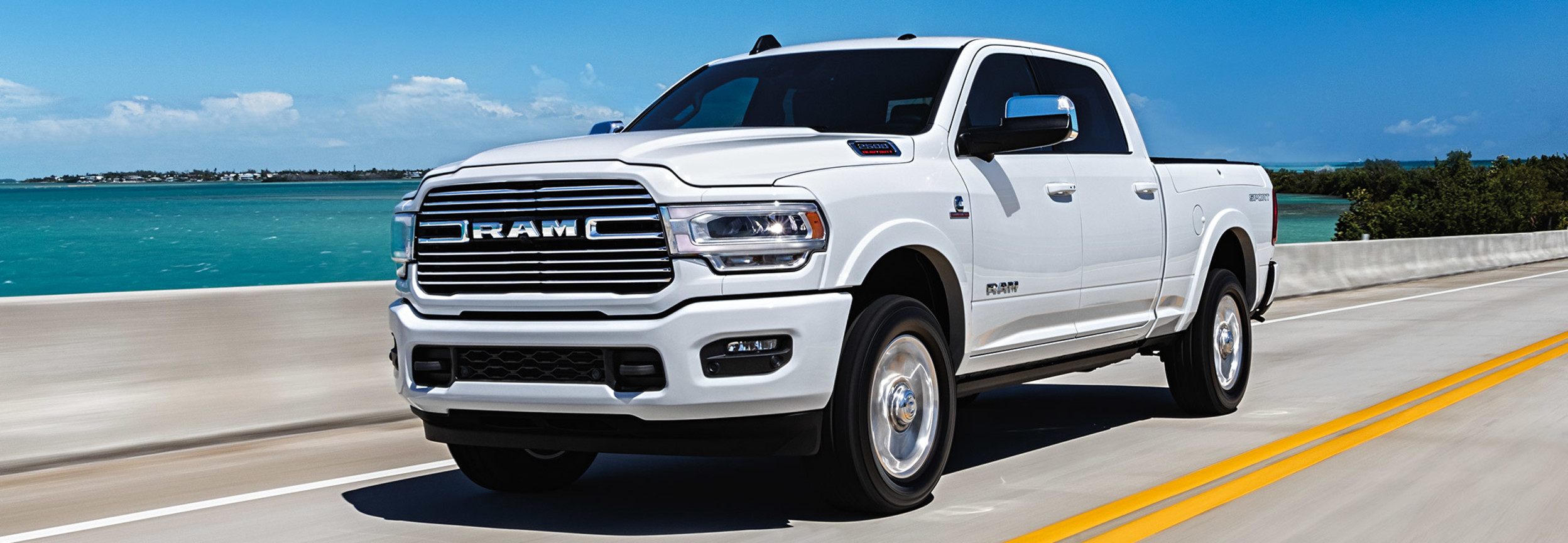 Front view of a white RAM 2500 driving down a road with the sea in the background