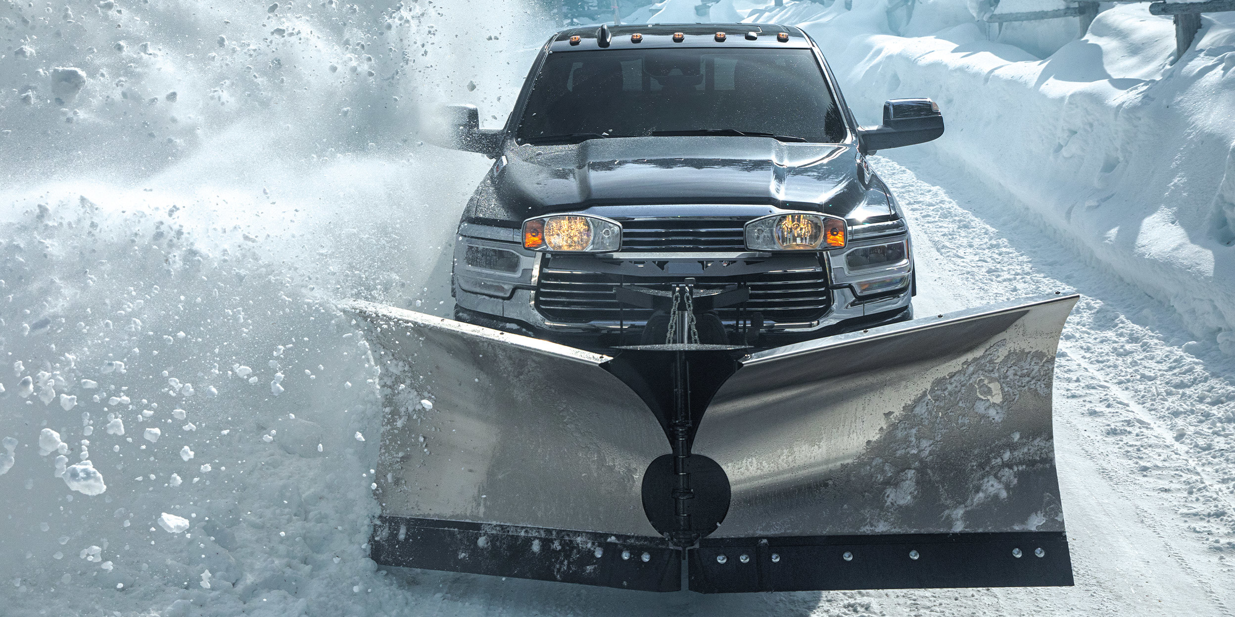 2500 Laramie in Maximum Steel with snow plows cleaning a snowy wooded road, headlights on.
