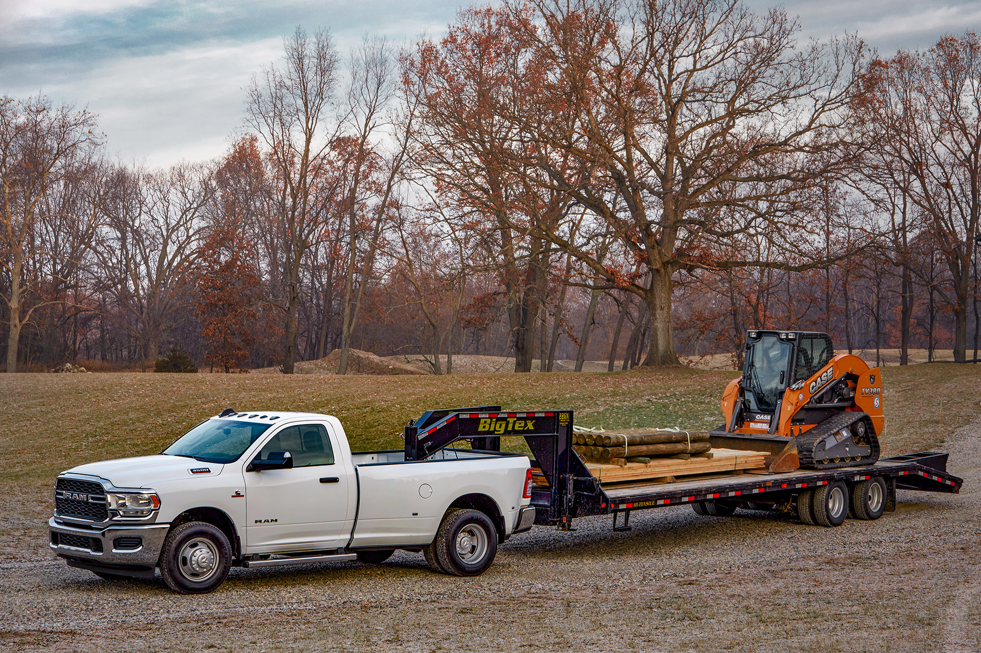 White 2020 Ram 3500 hauling a black trailer carrying wood and a skid steer loader.