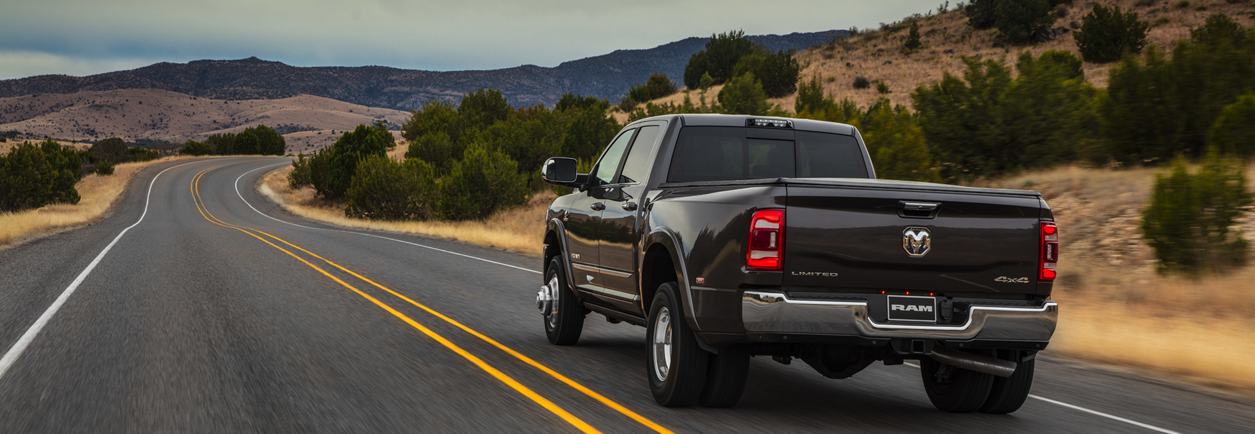 A rear view of a Diamond Black Crystal Pearl coloured 2021 Ram 3500 being driven on a road through a desert landscape.