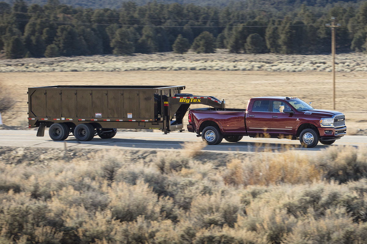 Un Ram 3500 2021 illustré en couche nacrée rouge tirant une grande remorque à benne sur la route, à côté d’un champ.
