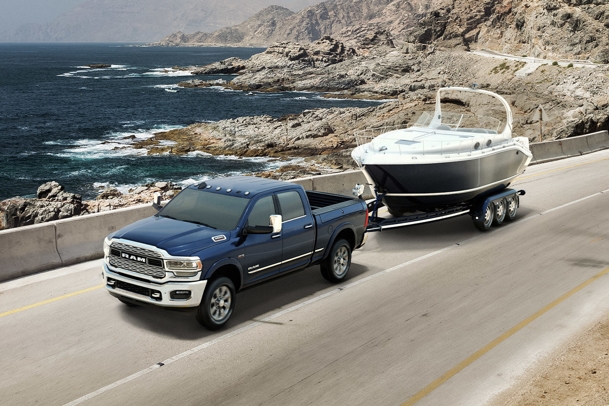 An elevated view of a Diamond Black Crystal Pearl coloured Ram 3500 towing a powerboat on a coastal road.