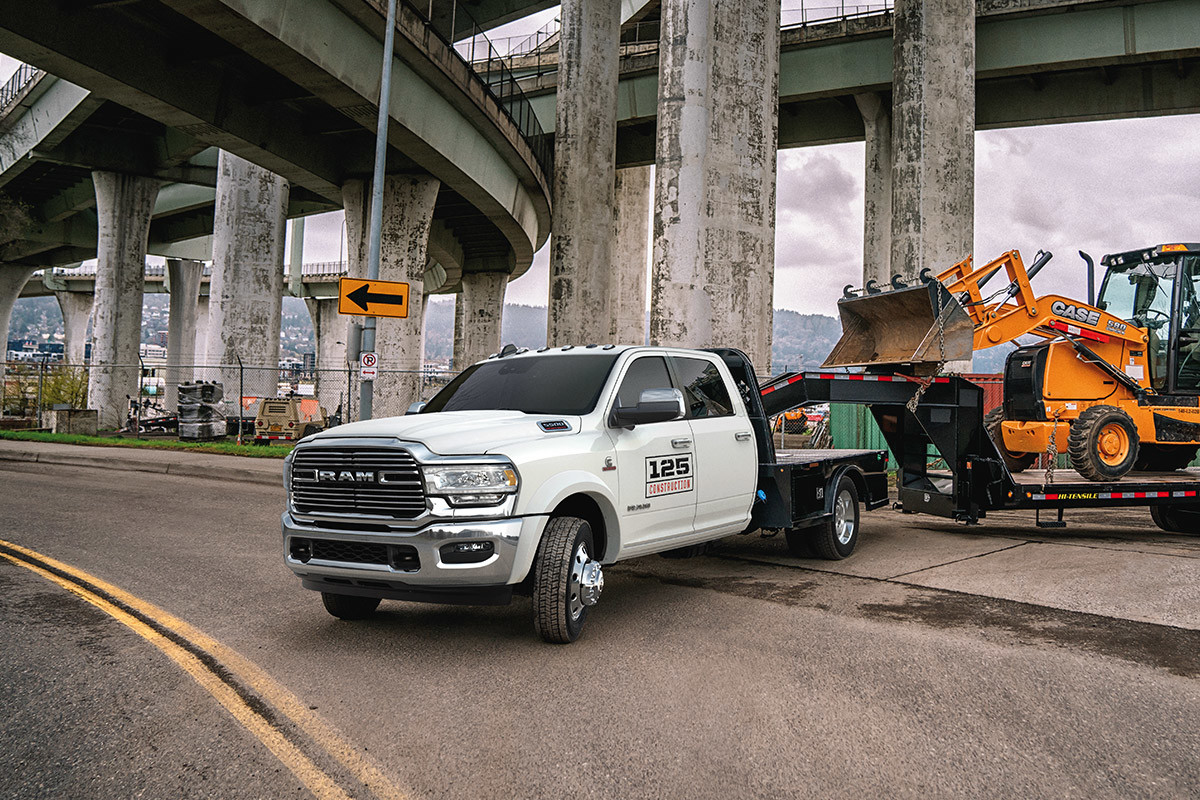 Un châssis-cabine Ram 2021 blanc éclatant tractant une chargeuse-pelleteuse sur une remorque sous un viaduc routier.
