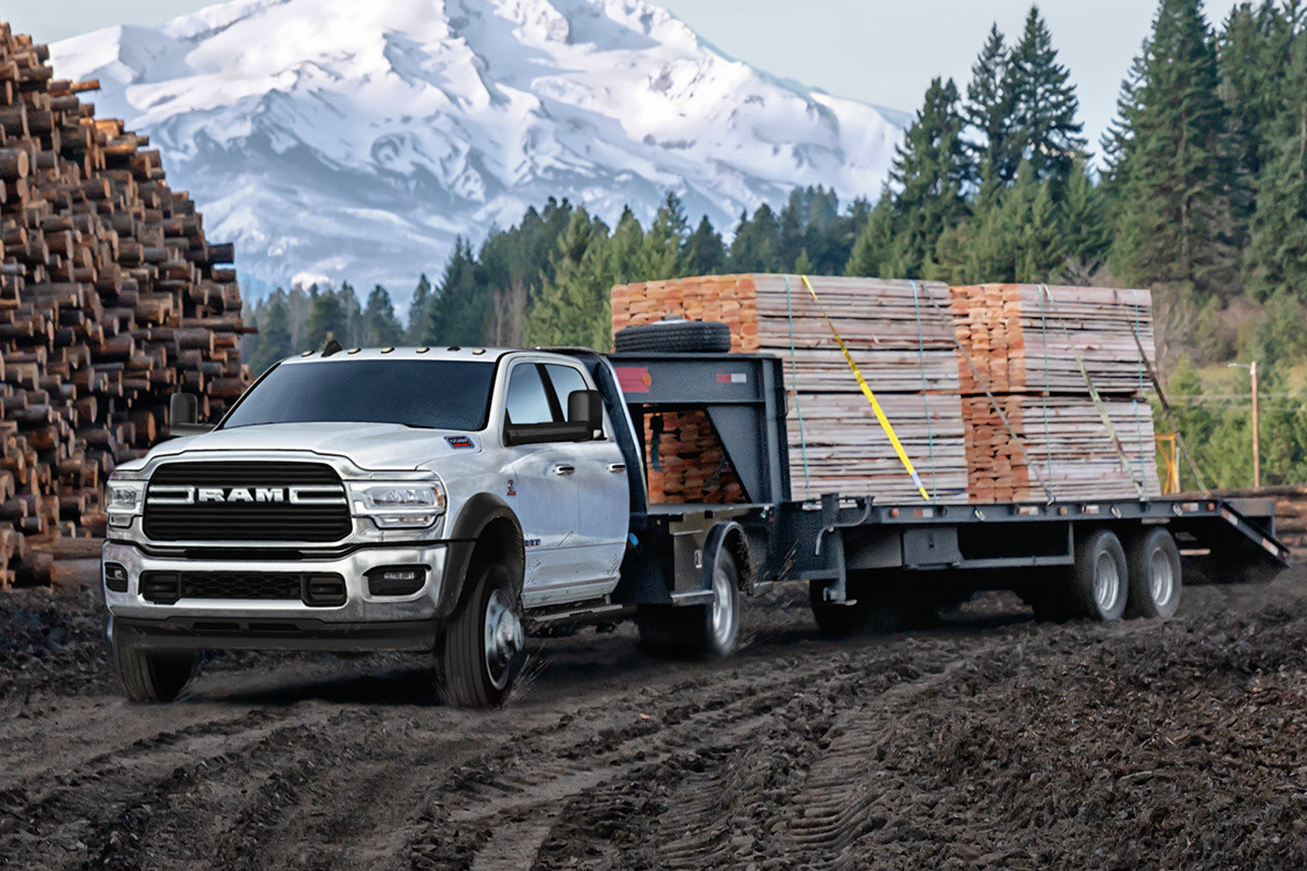 Vue avant d’un châssis-cabine Ram 2021 blanc éclatant tractant une remorque chargée de piles de bois sur une route de terre avec des montagnes enneigées au loin.