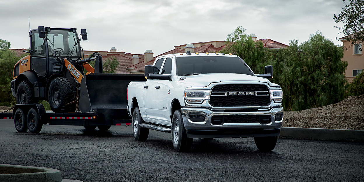 White Ram 2500 Heavy Duty pickup truck towing a skid steer loader on a road