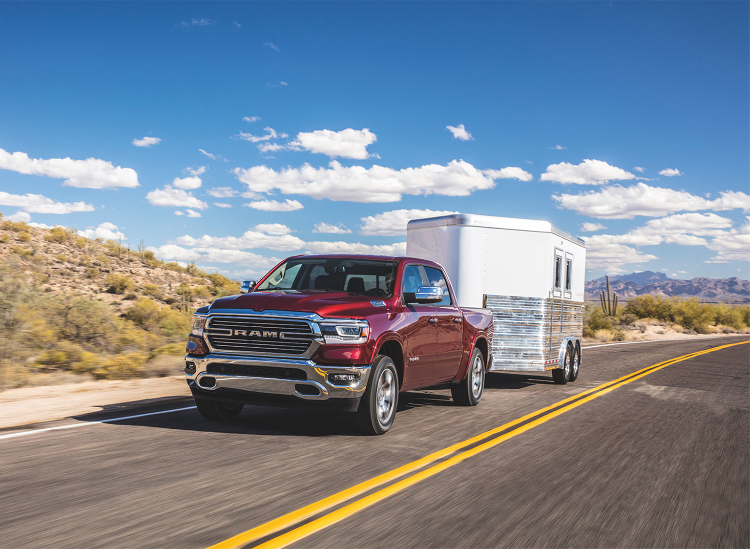 A red Ram 1500, towing a white trailer on a paved road through a desert area.