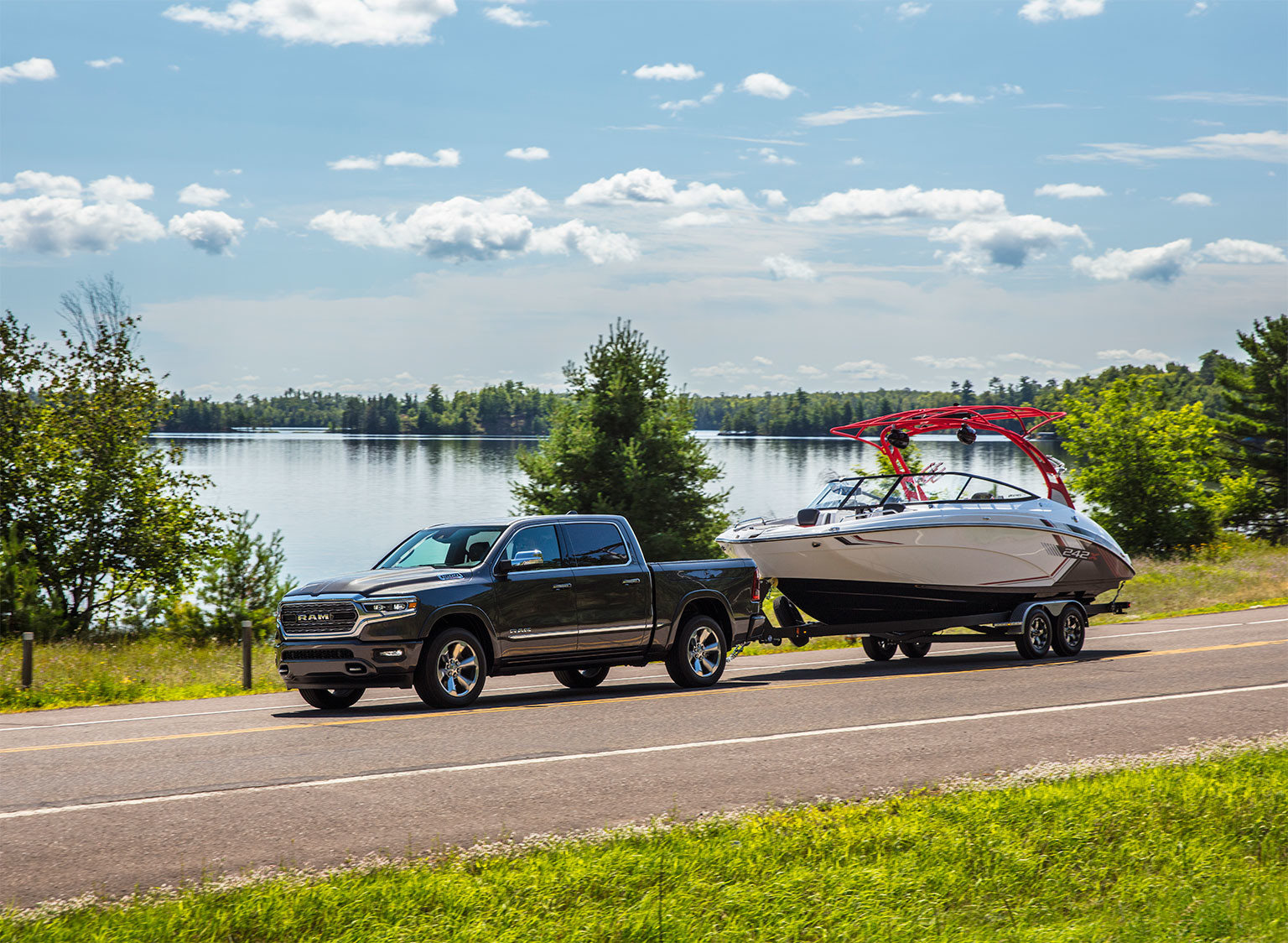 A grey Ram 1500 being driven on a paved road, towing a boat.