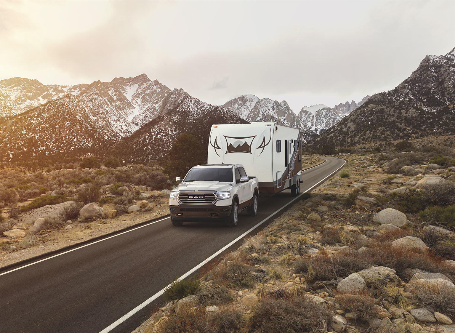 A white Ram 1500, towing a large travel trailer on a paved road through snowy mountains.