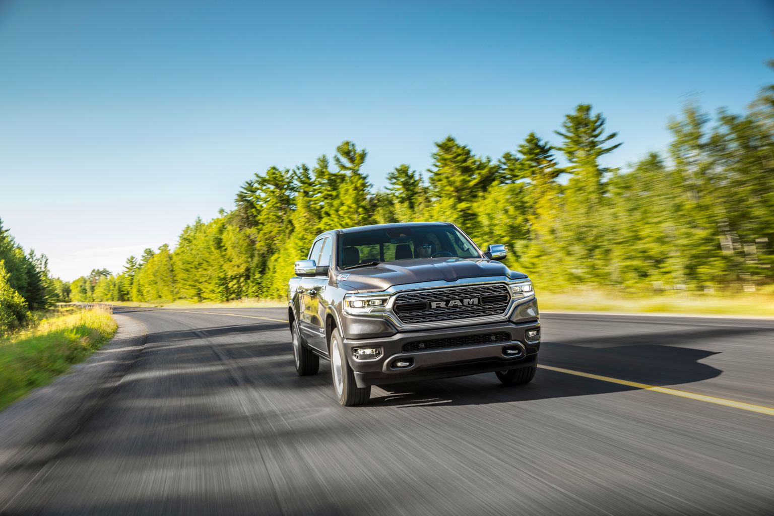 A grey Ram 1500 being driven on a paved road through tall trees.