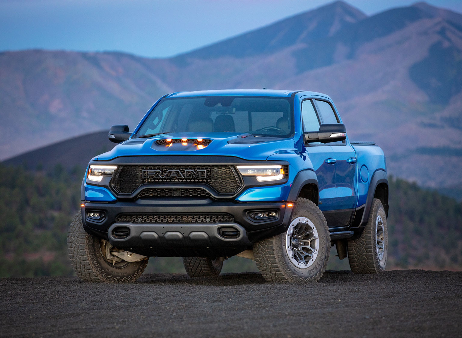 Front view of blue 2023 Ram 1500 TRX parked on a dirt road with mountains and trees in the background.