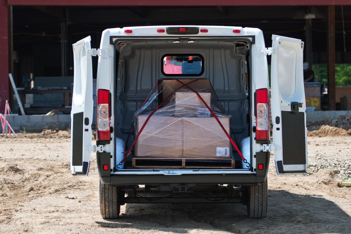Ram ProMaster stationné à proximité d’un chantier avec ses portes arrière ouvertes, laissant entrevoir des caisses empilées et arrimées à l’intérieur.