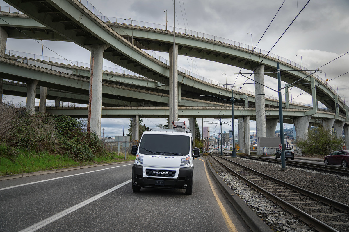 Ram ProMaster driving on a road, with a bridge and city buildings in the background.