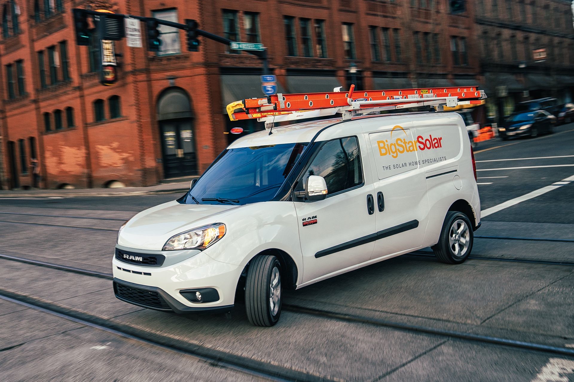 Front view of the white 2019 Ram ProMaster City with a ladder on its roof driving on a city street