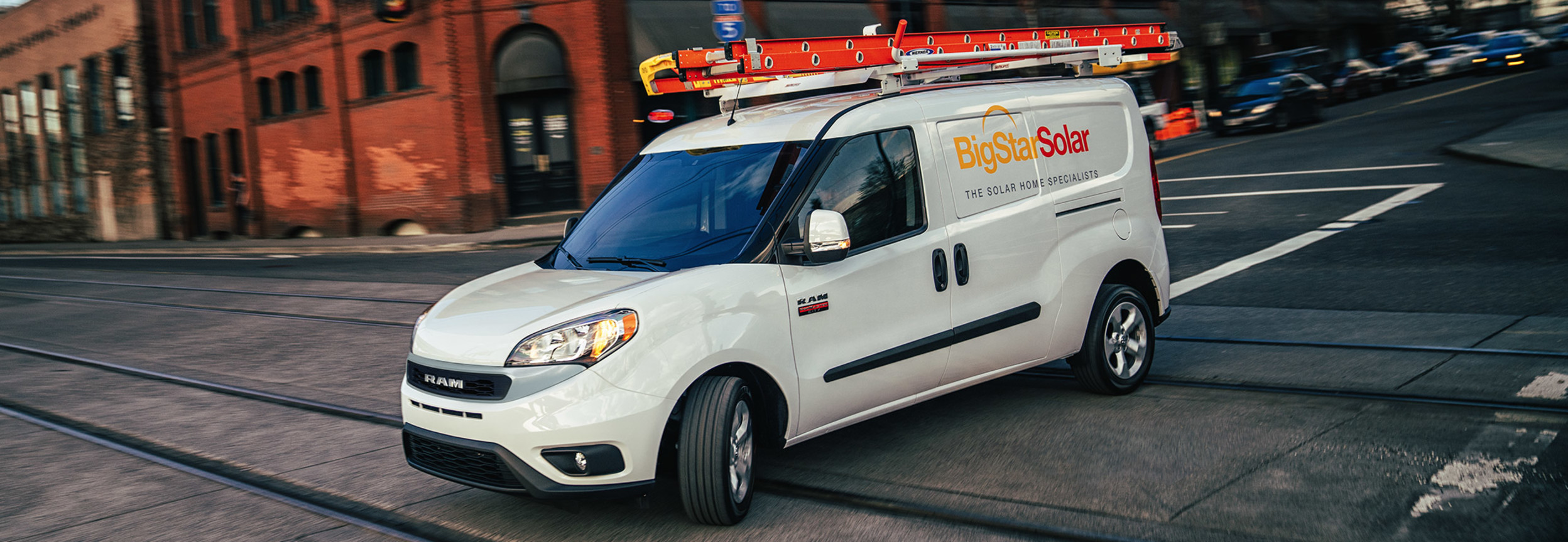 A white RAM ProMaster City with a ladder attached to its roof driving across a city road between traffic lights