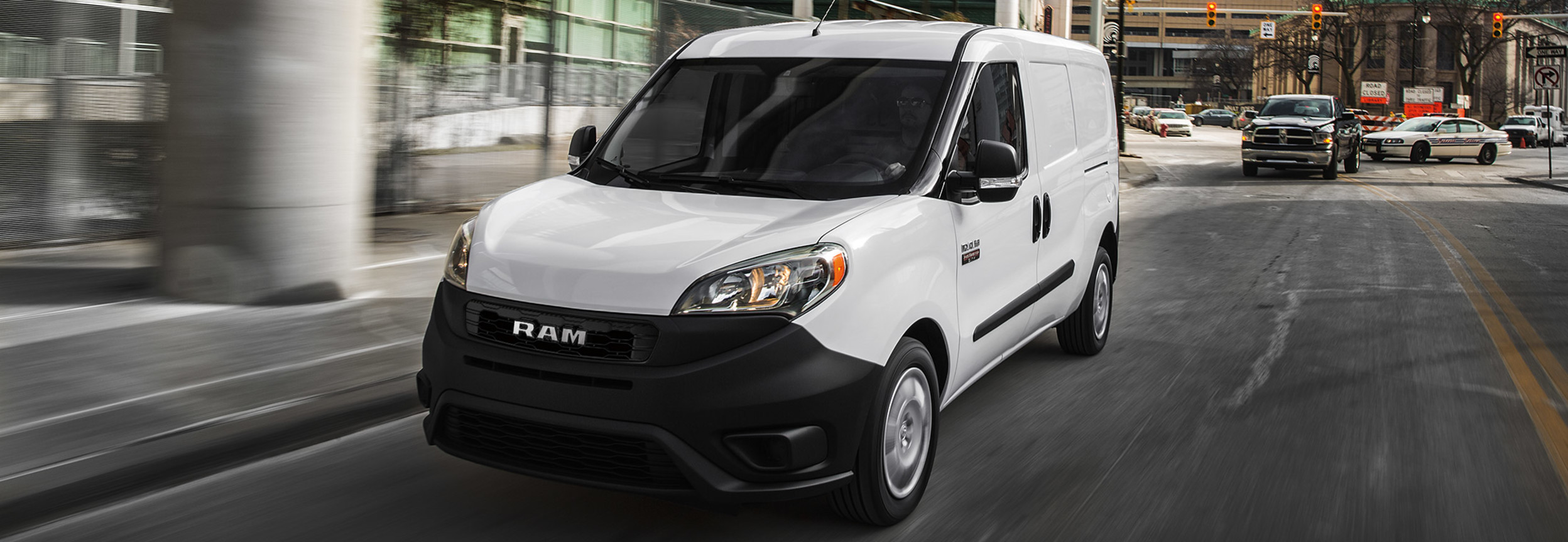 Un RAM Promaster City blanc roulant sous une autoroute sur une route de la ville