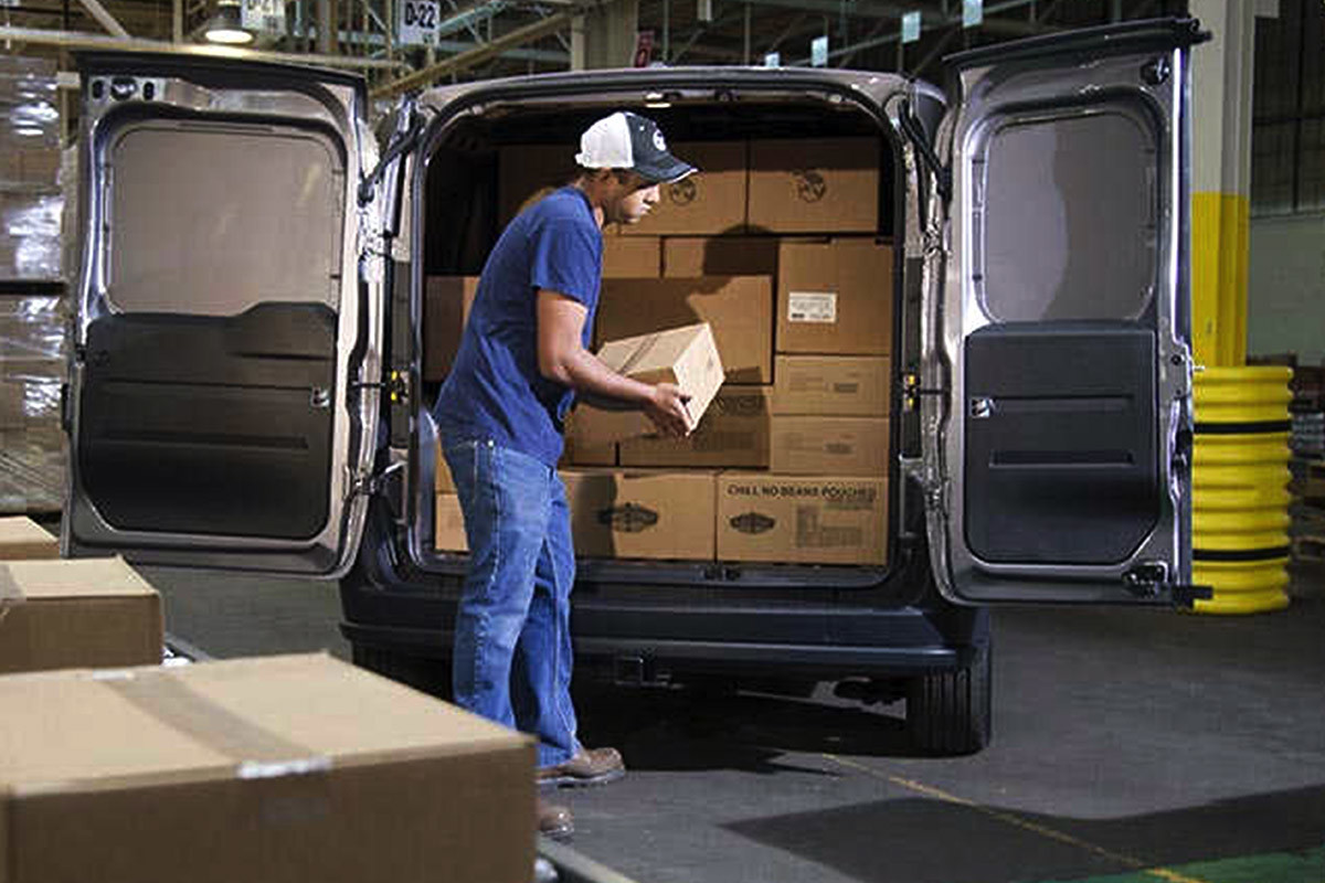 White 2021 Ram ProMaster City parked in a warehouse. Both rear doors are wide open and the cargo area is filled with packages. A worker is loading a package easily.