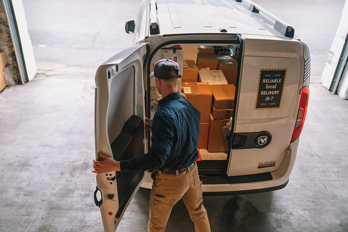 Ram ProMaster City 2021 blanc garé dans une aire de chargement. Un messager ouvre les portes arrière divisées 60/40 pour accéder facilement aux paquets qui se trouvent à l’intérieur.