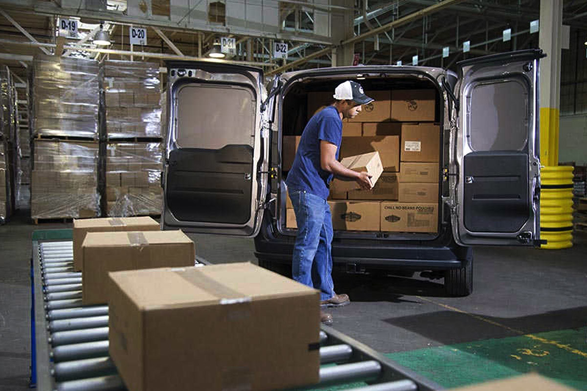 White 2021 Ram ProMaster City parked in a warehouse. Both rear doors are wide open, and the cargo area is filled with packages. A worker is loading a package easily.