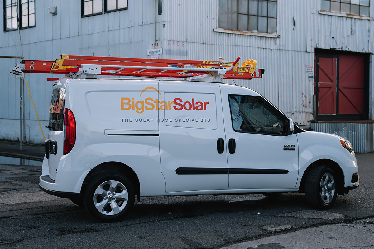White 2021 Ram ProMaster City customized with a ladder rack and business logo, parked in an industrial area.