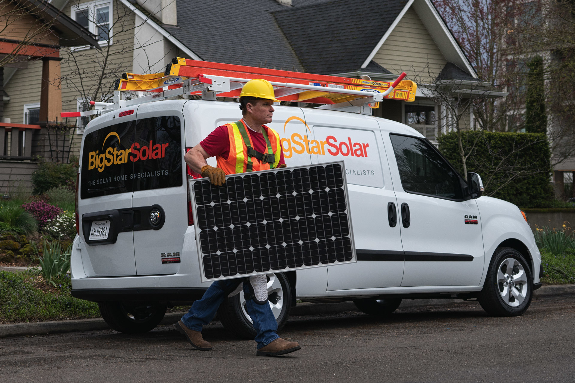 A repairman carries a solar panel from a white 2021 Ram ProMaster City parked in a residential neighborhood.
