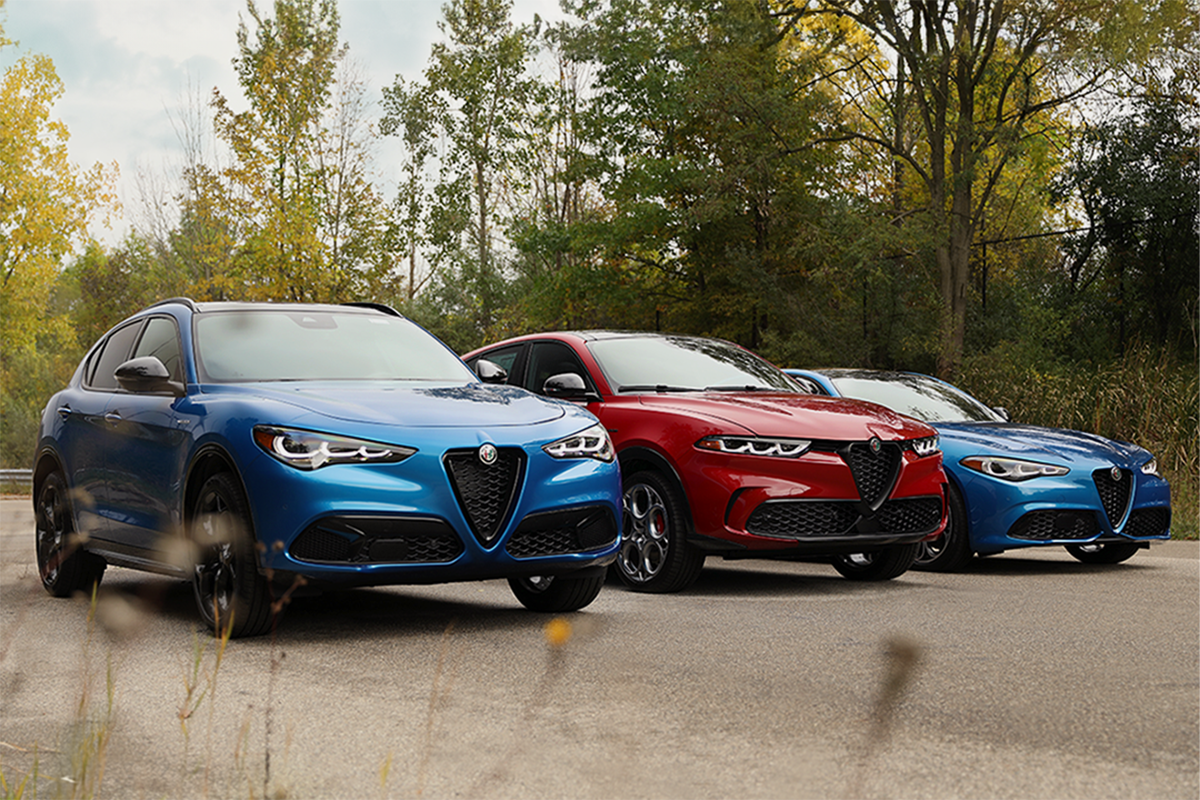 A Stelvio in blue, Tonale in red and a Guilia in blue parked on a dirt road with cloudy blue skies and trees visible behind.
