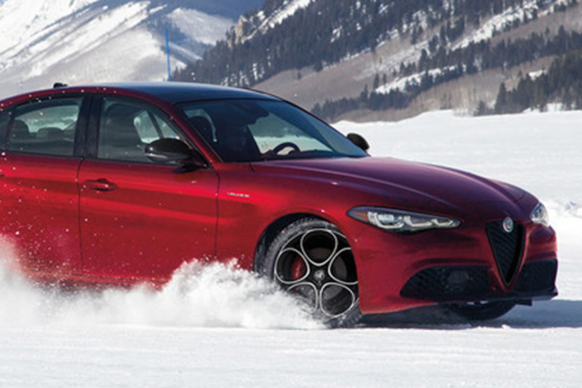Une Alfa Romeo Giulia rouge 2025 traversant un paysage enneigé avec de la neige pulvérisée autour des roues et des montagnes enneigées en arrière-plan.