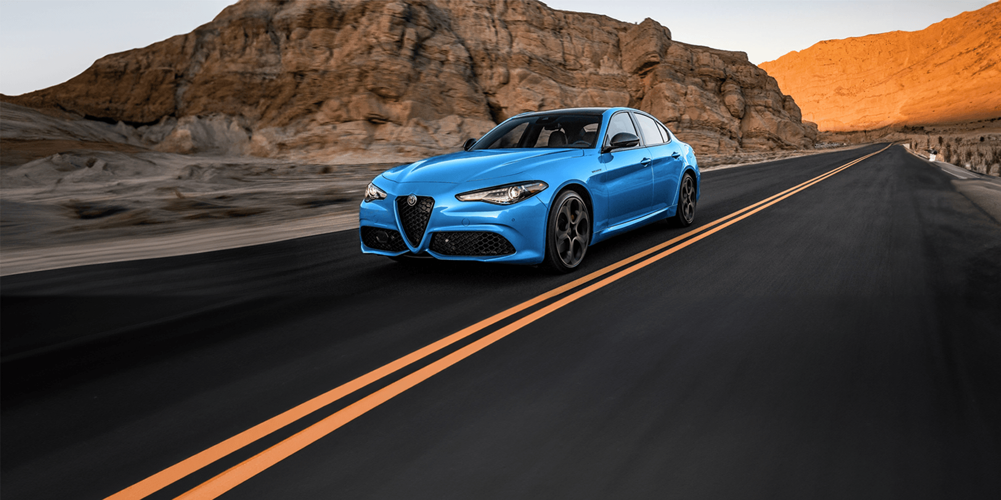 Une Alfa Romeo Giulia bleue 2025 roulant sur une route ouverte à travers un paysage désertique avec des formations rocheuses ensoleillées.