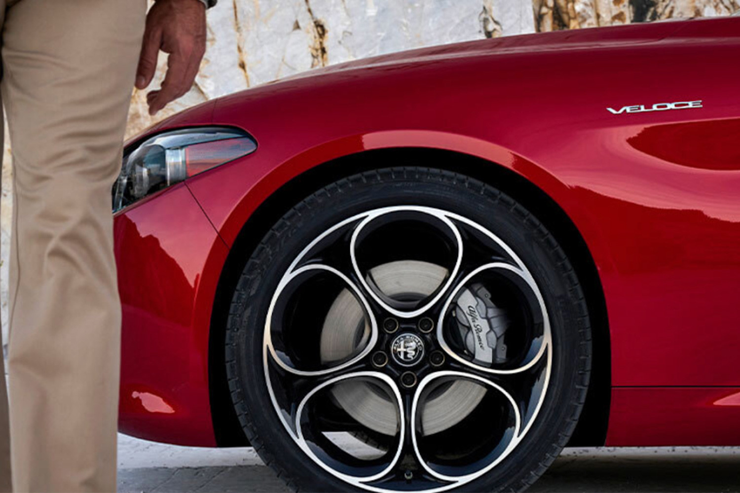 Vue partielle d'une Alfa Romeo rouge 2025, mettant l'accent sur le design des roues et le côté du véhicule. Une personne se tient à proximité sur un fond de roche texturée.