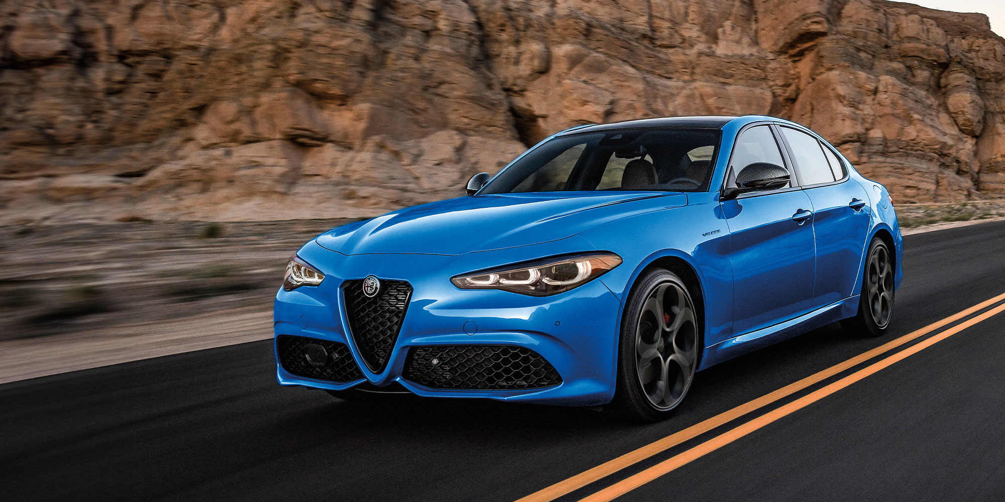 2025 Anodized Blue Metallic Alfa Romeo Giulia driving through a desert canyon with rocky cliffs and a clear sky at sunset.